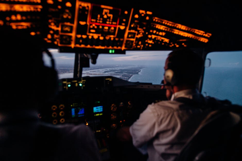 Back view of unrecognizable male pilots in headsets operating illuminating dashboard in cockpit while flying over sea in evening